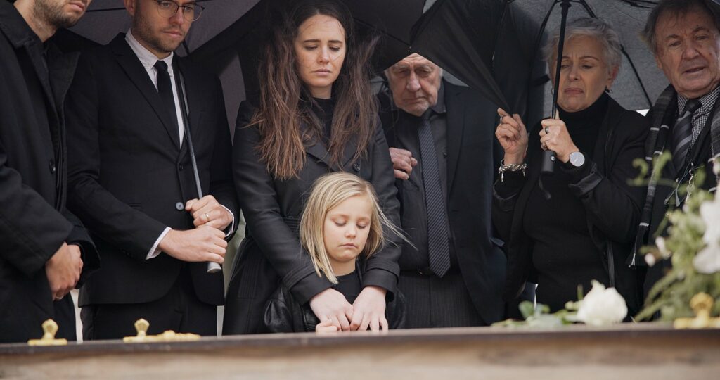 Loss, grief and people at funeral with umbrella, flowers and coffin, family with sad child at servi
