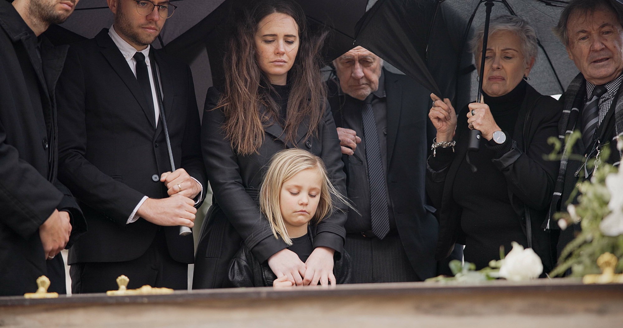 Loss, grief and people at funeral with umbrella, flowers and coffin, family with sad child at servi