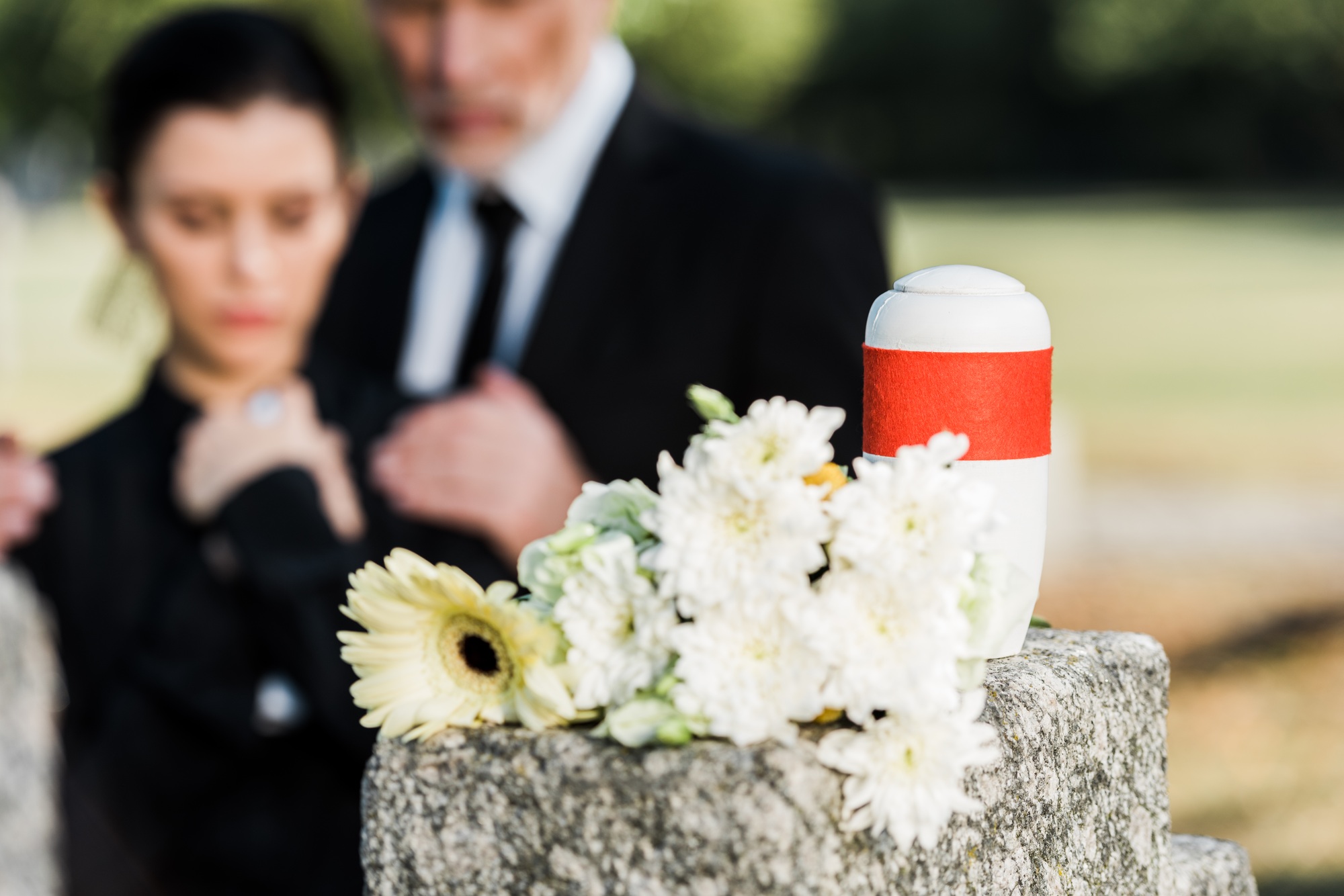 selective focus of white flowers and mortuary urn near man and woman in graveyard